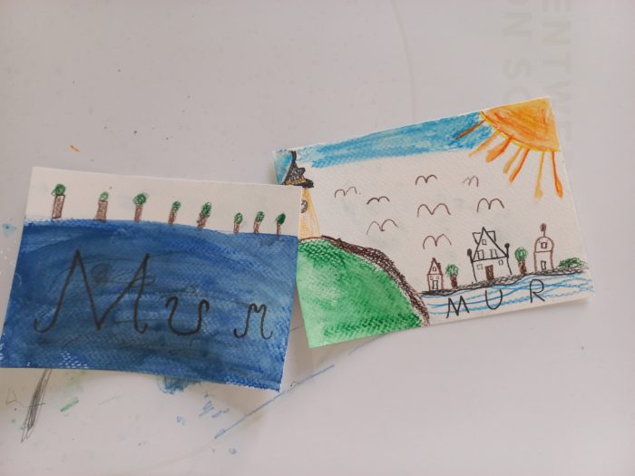 Auf einem weißen Tisch liegen zwei Kinderzeichnungen, die Ansichten der Mur zeigen.