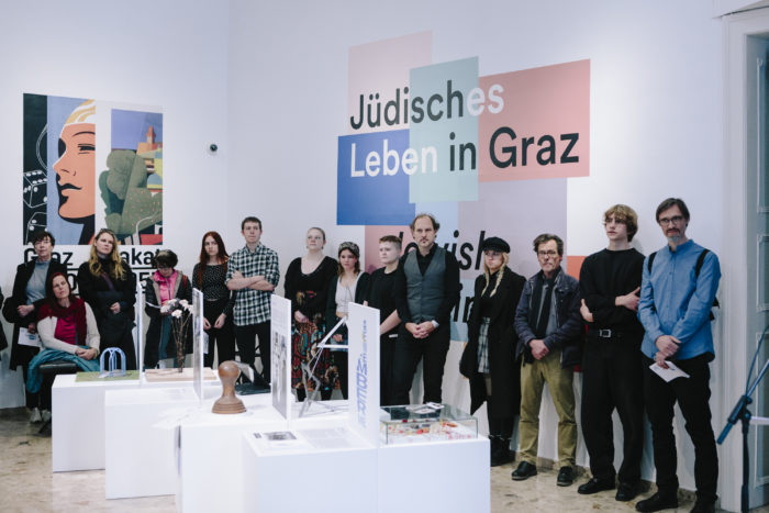 Im Foyer im ersten Stock des Graz Museums stehen Schüler*innen einer Klasse der Ortweinschule mit ihrem Lehrer vor weißn, quadratischen Podesten, auf denen von ihnen entworfene Modelle für Denkmäler ausgestellt sind.