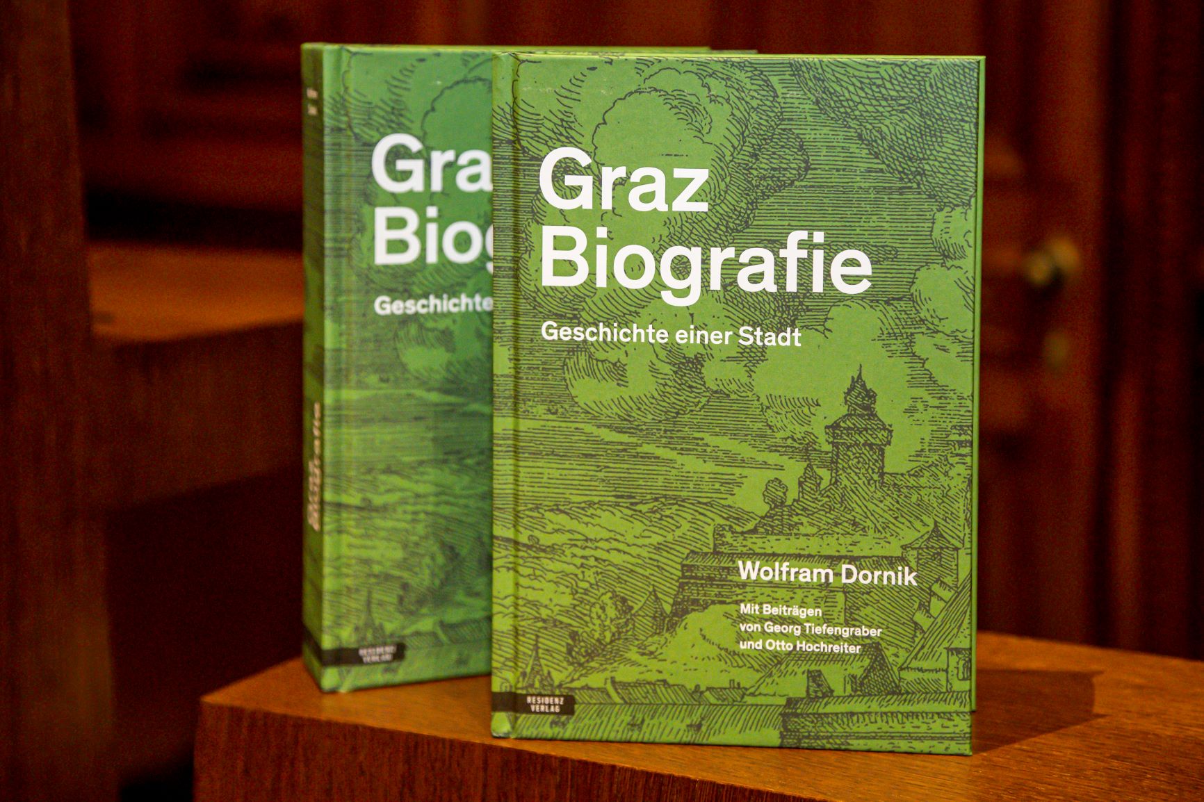 Auf einem dunkelbraunen Holzpult sind zwei exemplare de Buches Graz Biografie nebeneinander aufgestellt.