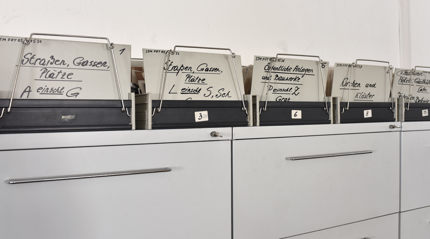 Auf einem hellgrauen Aktenschrank stehen Kistchen mit historischen Fotografien von Graz.