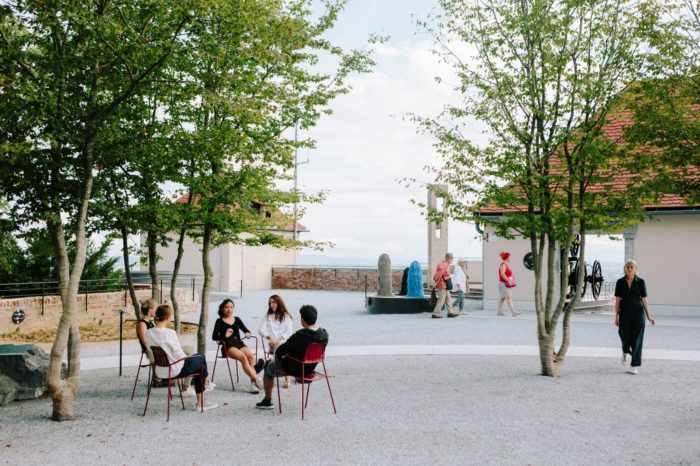 Im Innenhof des Graz Museums Schlossberg sitzen vier Personen unter Bäumen auf Sesseln, im Hintergrund sind zwei Personen zu sehen, die im Hof herumgehen.
