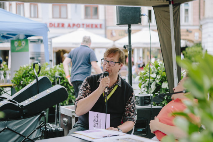 Ein Mann mit Brille sitzt am Grazer Hauptplatz auf einem Stuhl und spricht in einen Mikrofon, das er in der Hand hält.