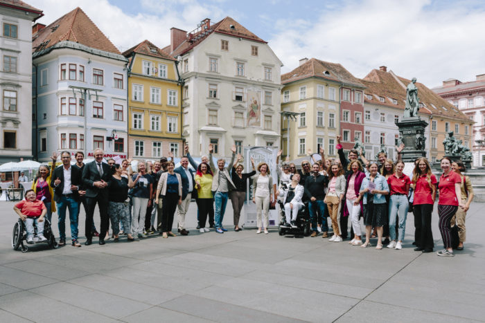 Auf einem Gruppenfoto stehen etwa 30 Personen in einem Halbkreis auf dem Grazer Hauptplatz. Im Hintergrund sind Häuser zu sehen.