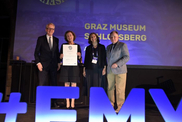 Otto Hochreiter, Sibylle, Dienesch und Martina zerovnik stehen mit einem Juror auf der Büühne und lachen. Dienesch hält eine Urkunde in der Hand.