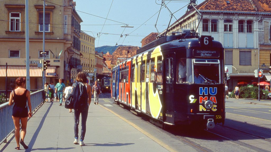 Auf der Erzherzog-Johann-Brücke gehen Menschen, zwei sind unterwegs in Richtung Südtirolerplatz. Eine Straßen der Linie 6 mit einer Reklame von HUMANIC fährt über die Brücke in Richtung Hauptplatz. Im Hintergrund ist das ehemalige Eiserne Haus zu sehen.