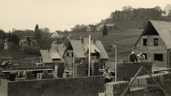 Schwarzweiß-Foto mit Bauarbeitern, die gerade an einem neuem Haus am Viktor-Geram-Weg arbieren. BIsher sind nur Teile von Ziegel-Wänden des Hauses aufgebaut.