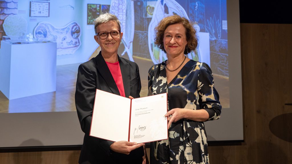Sektionschefin Theresia Niedermüller und Direktorin Sibylle Dienesch stehen nebeneinander und halten eine Urkunde in ihren Händen. Beide lächeln.